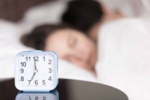 A blue alarm clock displaying 11:12 sits on a bedside table, with a blurred figure reclining in bed behind it.