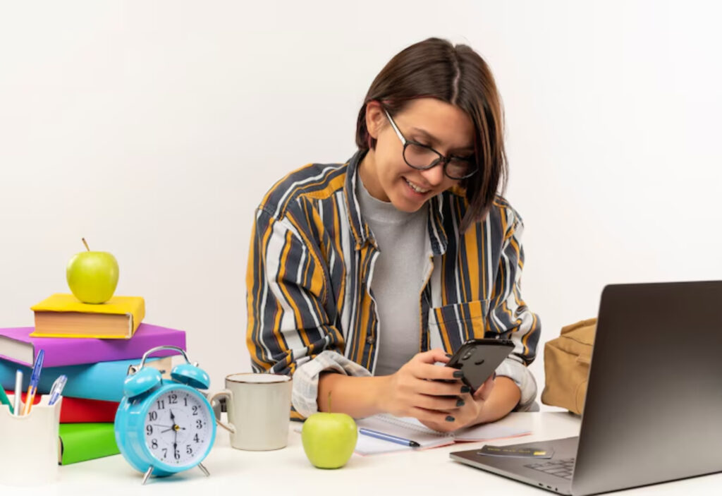 A person in a striped shirt sits at a desk with a laptop, books, clock, mug, and green apples, focused on their smartphone.