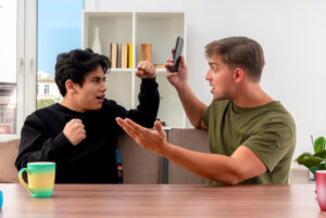 Two people are engaged in a heated discussion at a table with colorful mugs, one gesturing emphatically while holding a smartphone.