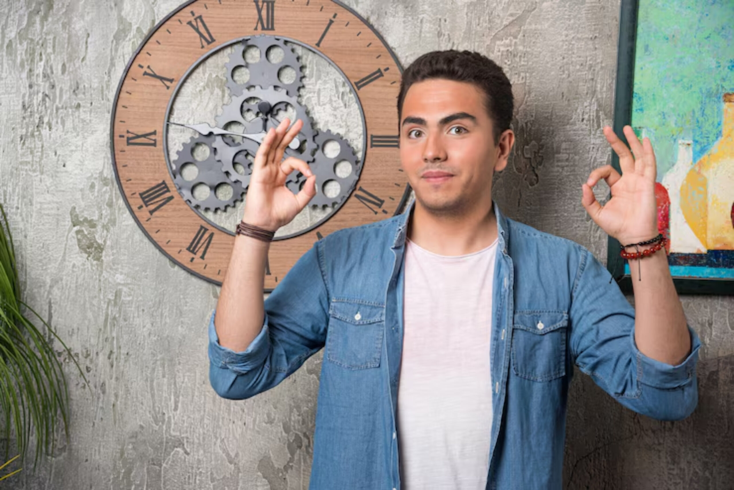 A person in a denim shirt is posing with hand gestures in front of a decorative wall clock and a colorful painting.