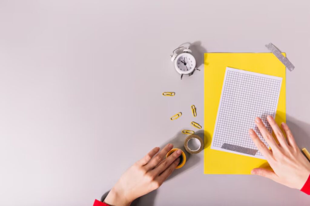 A flat lay of a workspace with a hand on yellow graph paper, surrounded by a clock, yellow paperclips, washi tape, and a bright yellow folder. The mood is organized and creative.