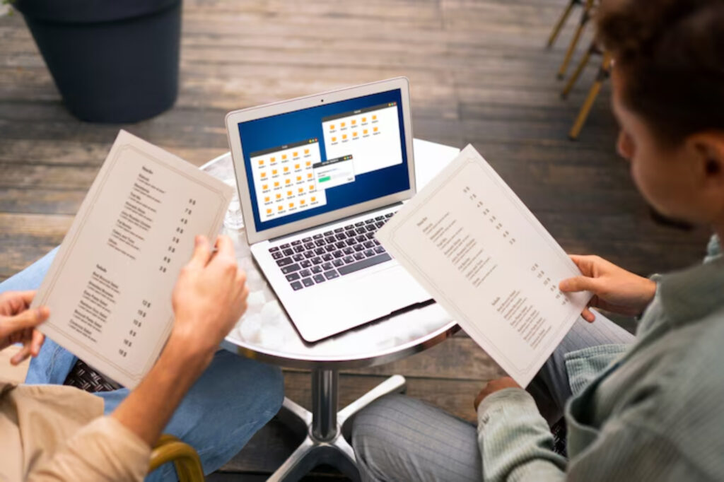 Two individuals sit at a round table, reviewing printed menus while a laptop displays open folders on its screen.