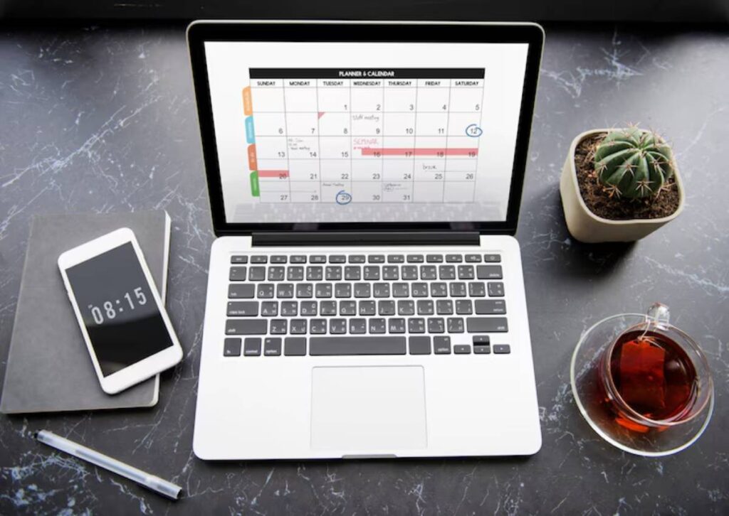 A laptop displaying a colorful calendar sits on a marble desk alongside a smartphone, notebook, pen, a cactus, and a cup of tea.