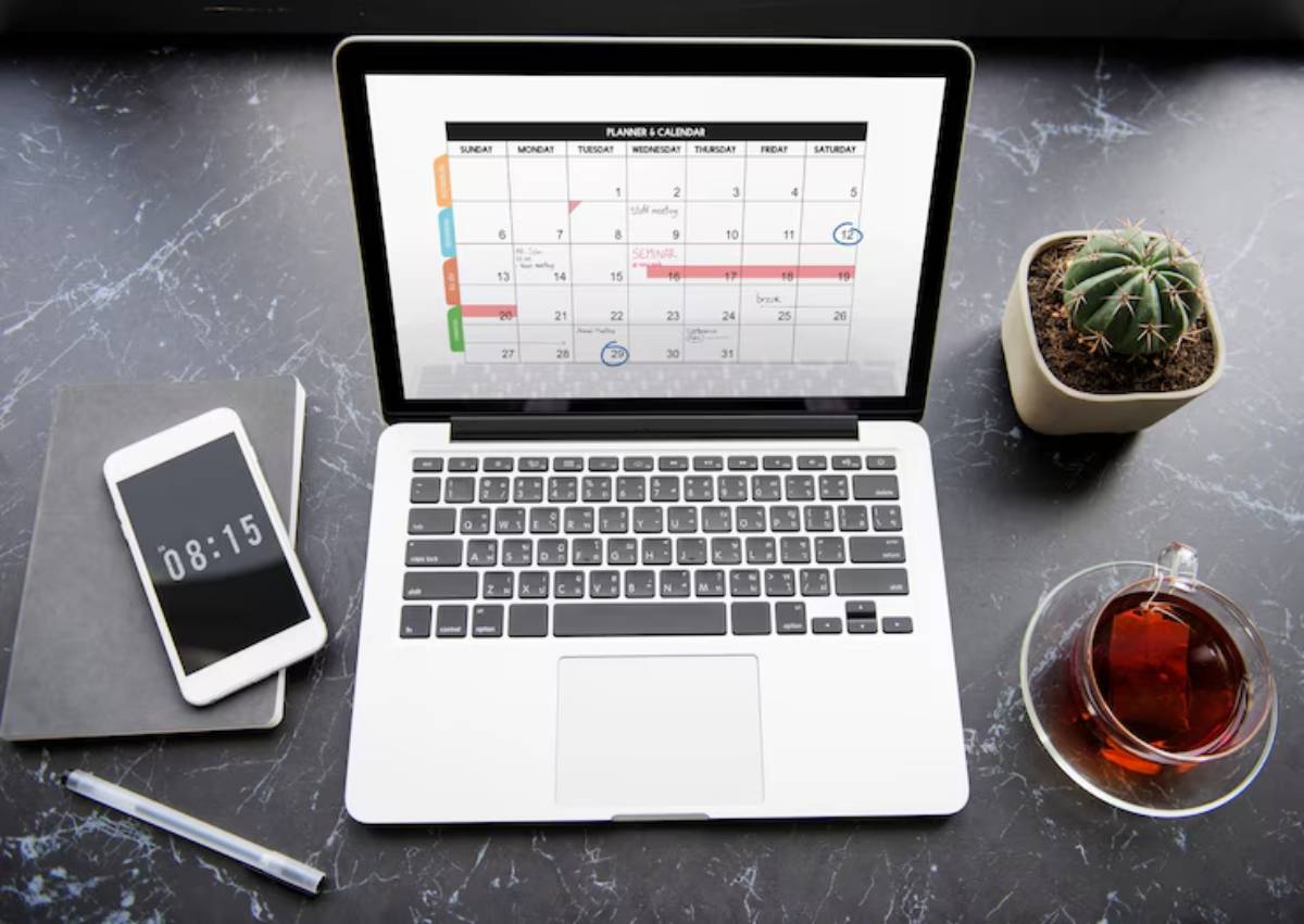A laptop displaying a colorful calendar sits on a marble desk alongside a smartphone, notebook, pen, a cactus, and a cup of tea.