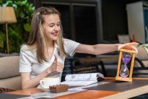 Smiling woman giving a thumbs-up while holding a wooden hourglass with purple sand. She sits at a table with an open book, coffee, and chocolate bar.