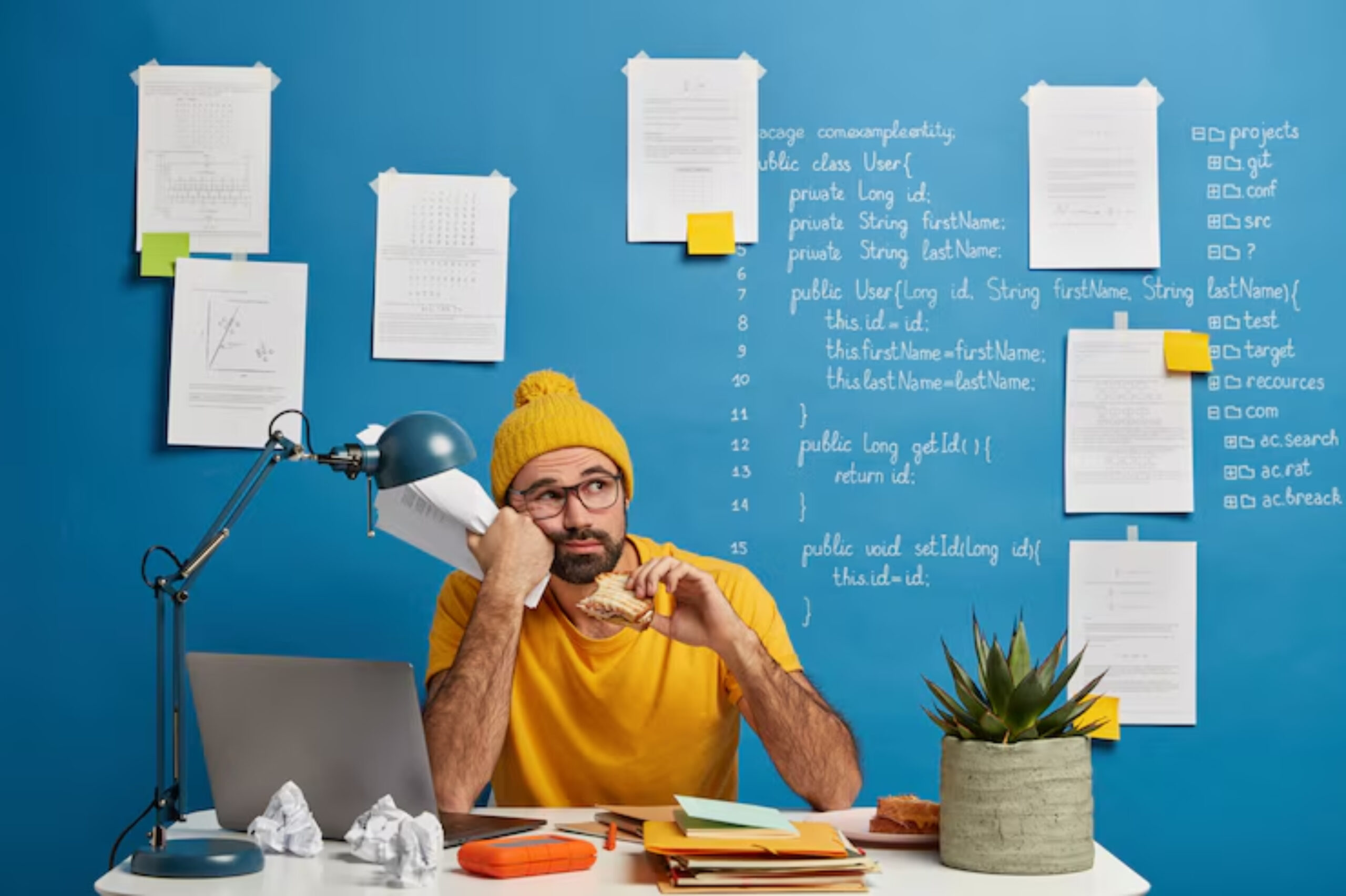 A person in a yellow beanie sits at a desk with a laptop, surrounded by papers and a potted plant, against a blue wall filled with notes.