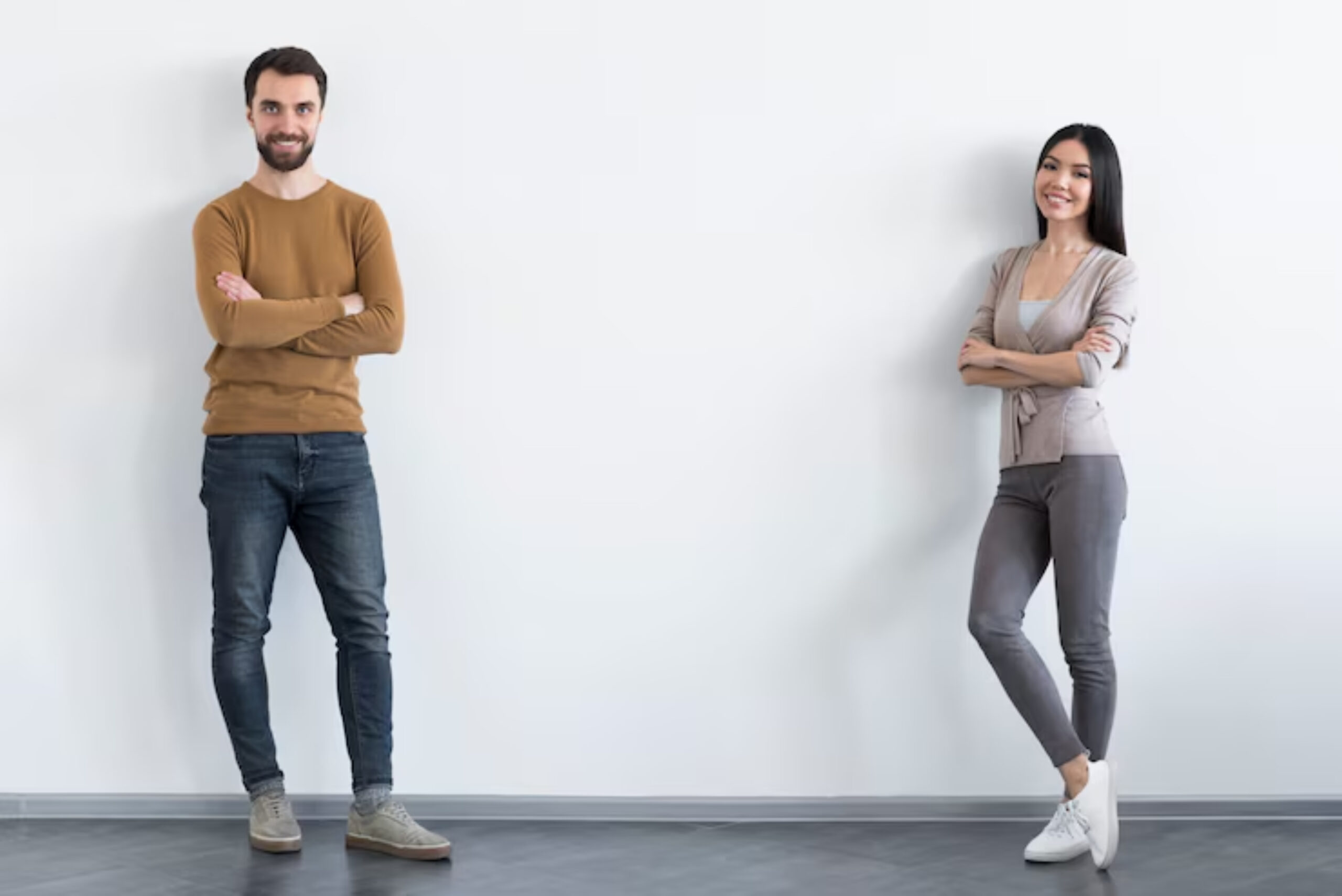 Two individuals stand on opposite sides of a blank wall, arms crossed, in casual attire. The space is bright and minimalistic.