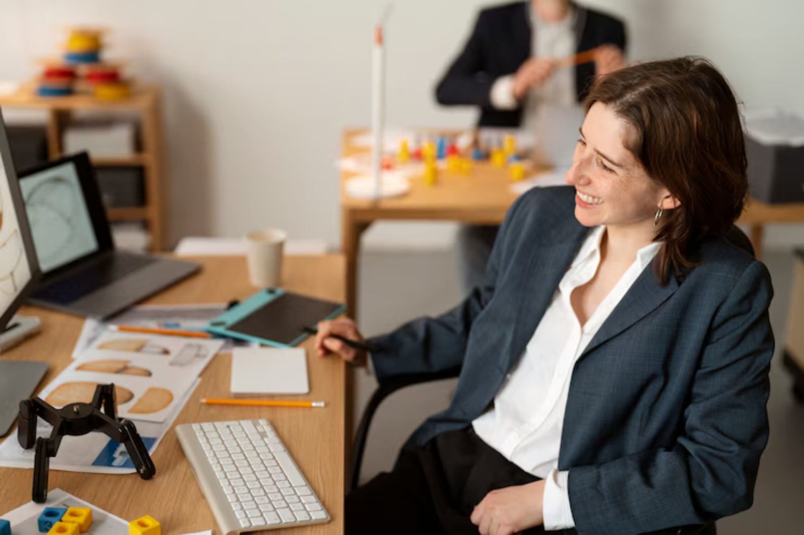 A woman in a blazer sits at a desk with various design materials, a laptop, and colorful building blocks, focused on her work.