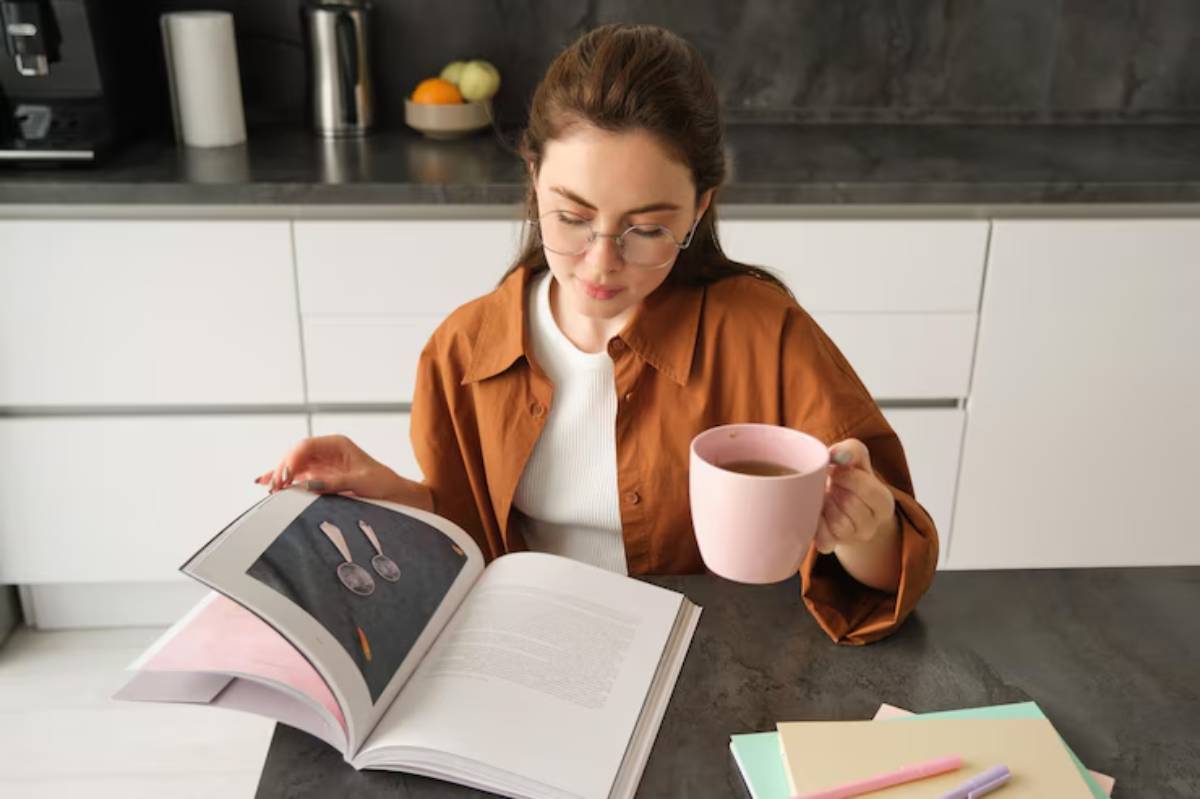 A person in an orange shirt sits at a kitchen table holding a pink mug, looking at a large cookbook open to a page with utensils.