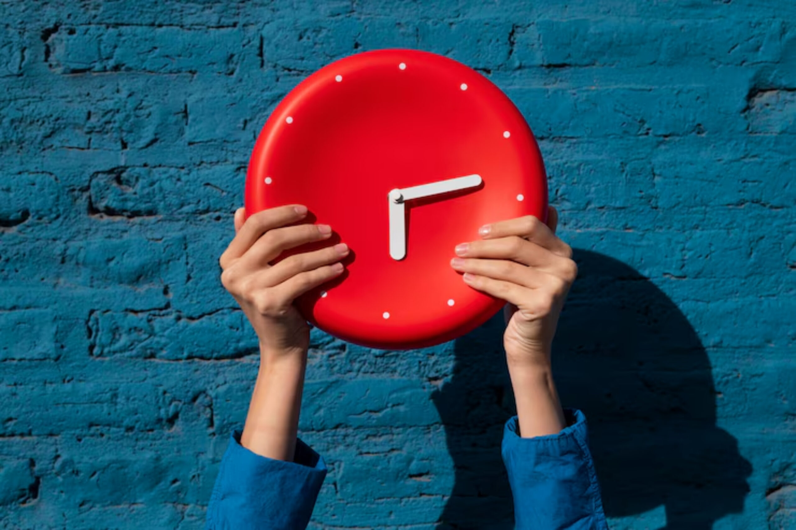 A pair of hands holds a bright red wall clock against a textured blue wall. The clock shows 1 o'clock.