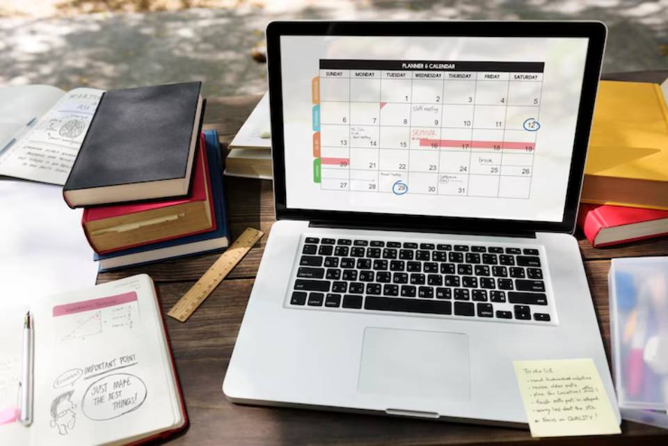 A laptop displaying a colorful calendar sits on a wooden table surrounded by books, a ruler, and notebook planners.