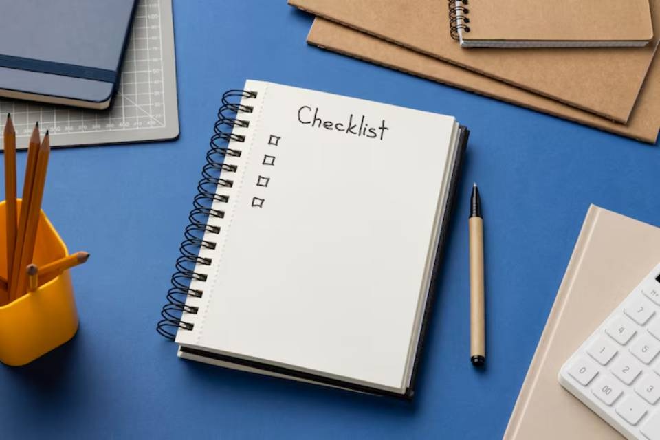 A blank checklist notebook on a blue desk, surrounded by stationery including a pen, notepads, and a pencil holder.