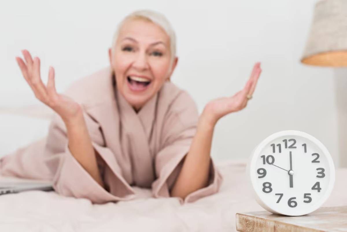 A person in a soft pink robe lies on a bed, gesturing expressively towards a white alarm clock displaying 10:10.