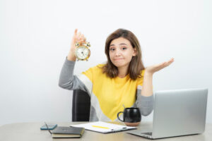 A person in a yellow and gray outfit holds a vintage clock while sitting at a desk with a laptop, notebook, and coffee mug.