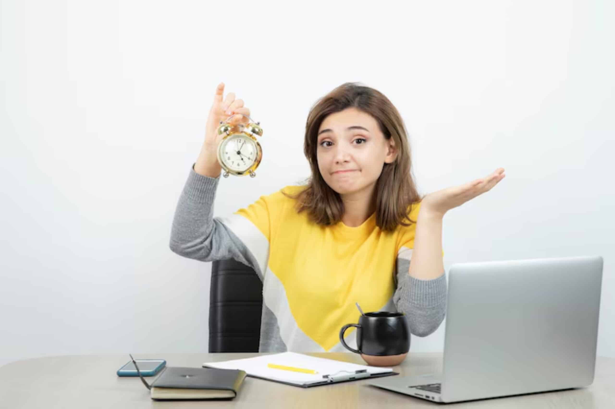 A person in a yellow and gray outfit holds a vintage clock while sitting at a desk with a laptop, notebook, and coffee mug.