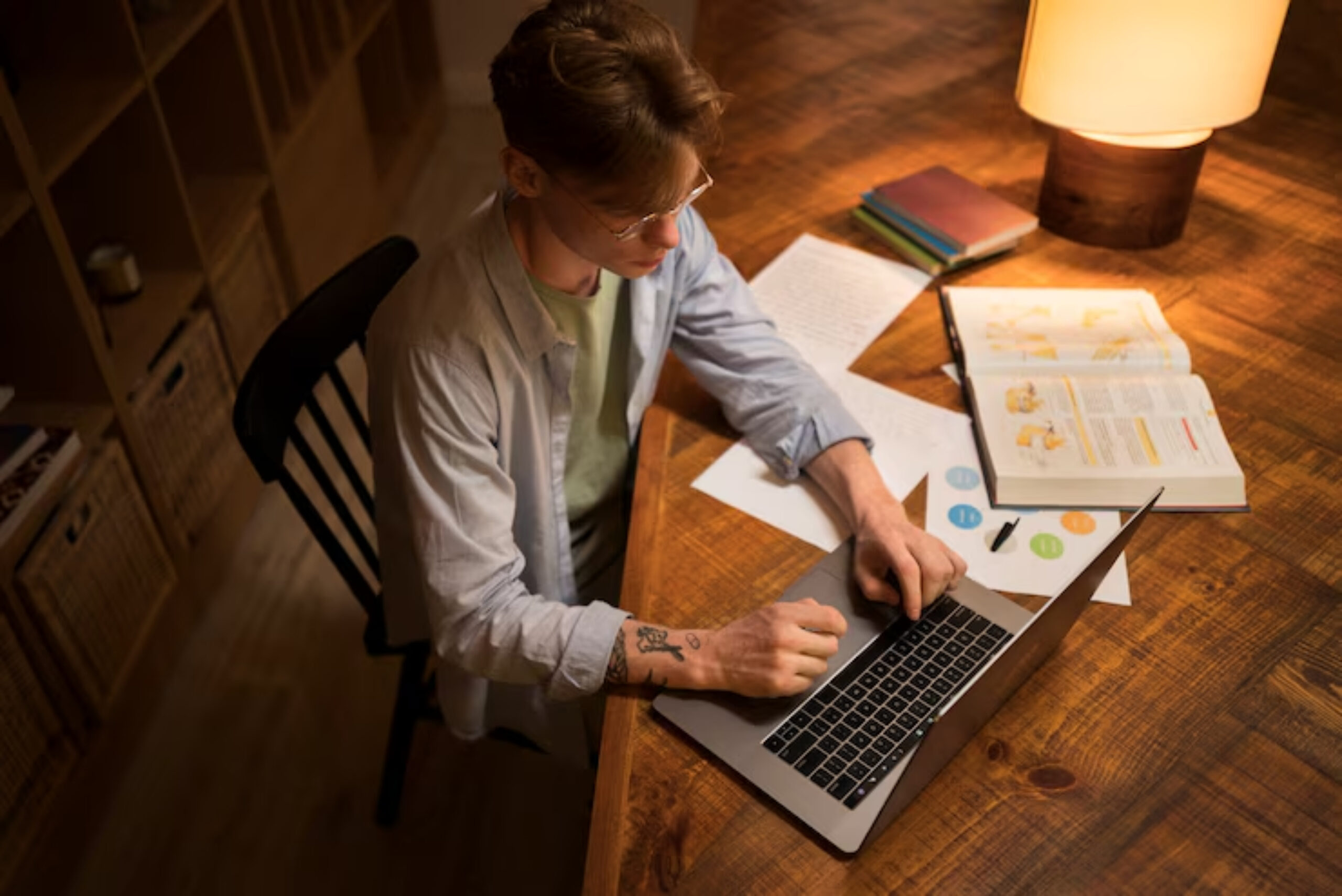 A person works on a laptop at a wooden desk covered with papers and books under soft, warm lighting.