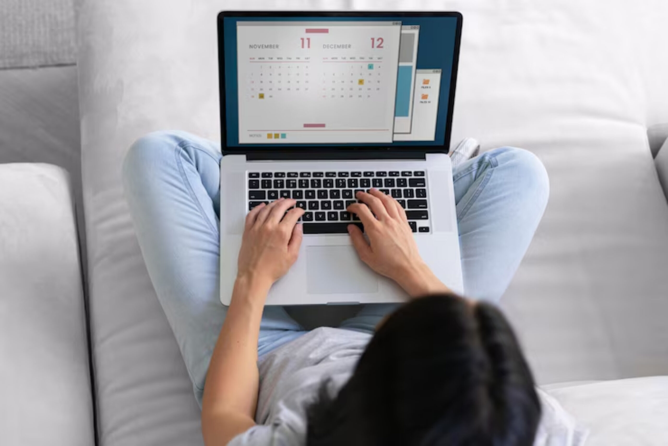 A person types on a laptop displaying a calendar, seated on a couch with light blue jeans and a casual t-shirt.