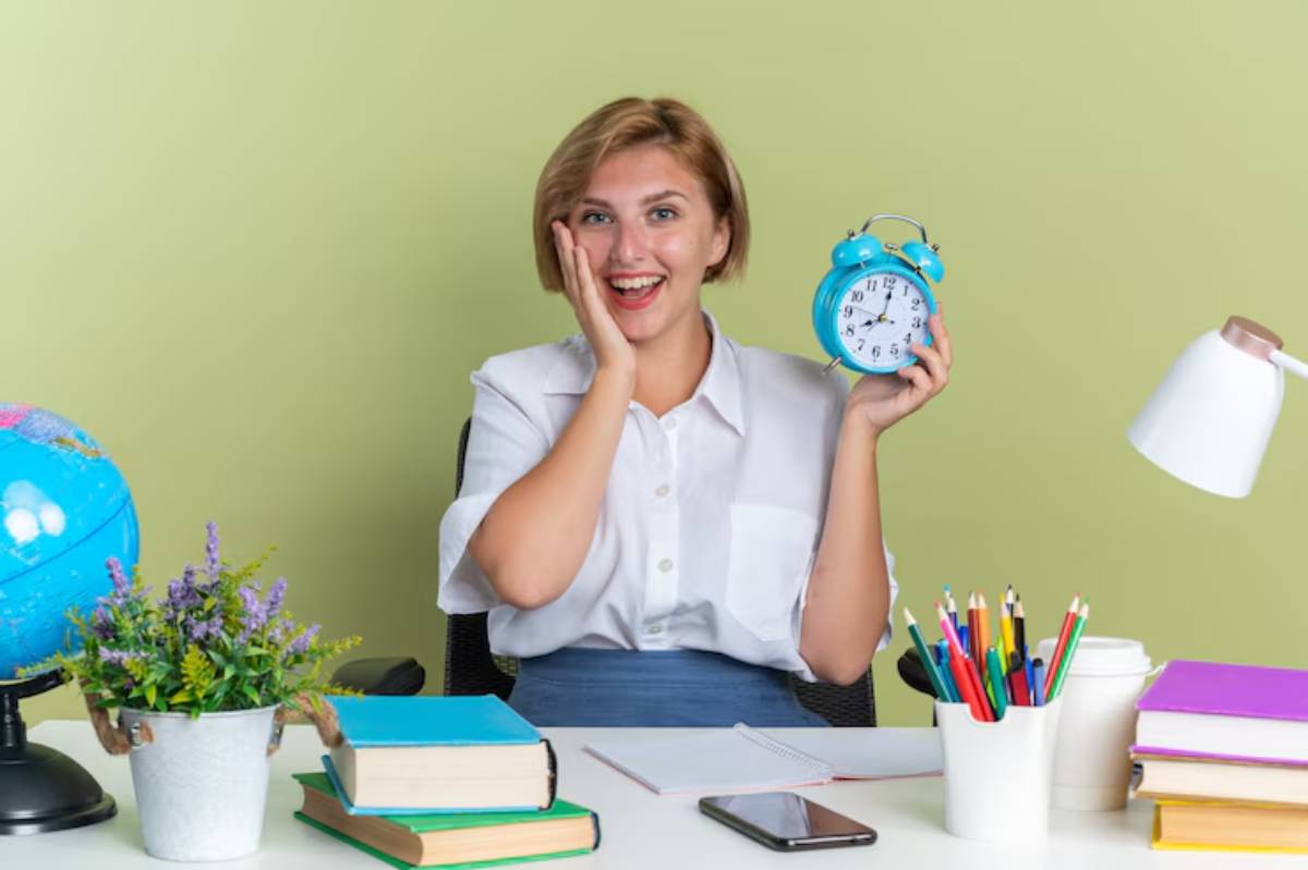 A person holding a blue alarm clock sits at a desk cluttered with books, stationery, and a globe, against a light green wall.