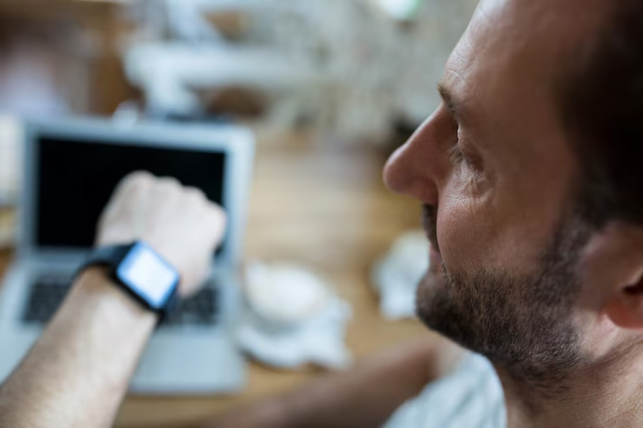 A person's wrist wearing a smartwatch is raised towards a laptop with a blank screen, on a wooden table alongside a coffee cup.