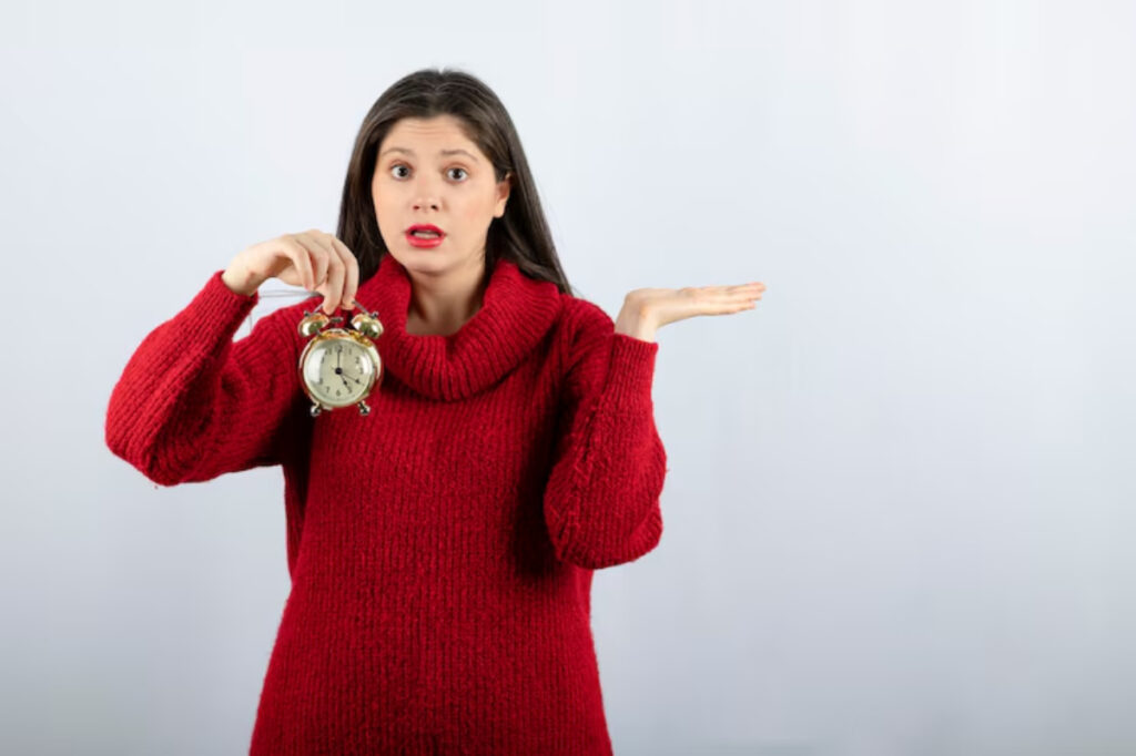 A person in a cozy red sweater holds a vintage alarm clock in one hand while raising the other hand in a questioning gesture.