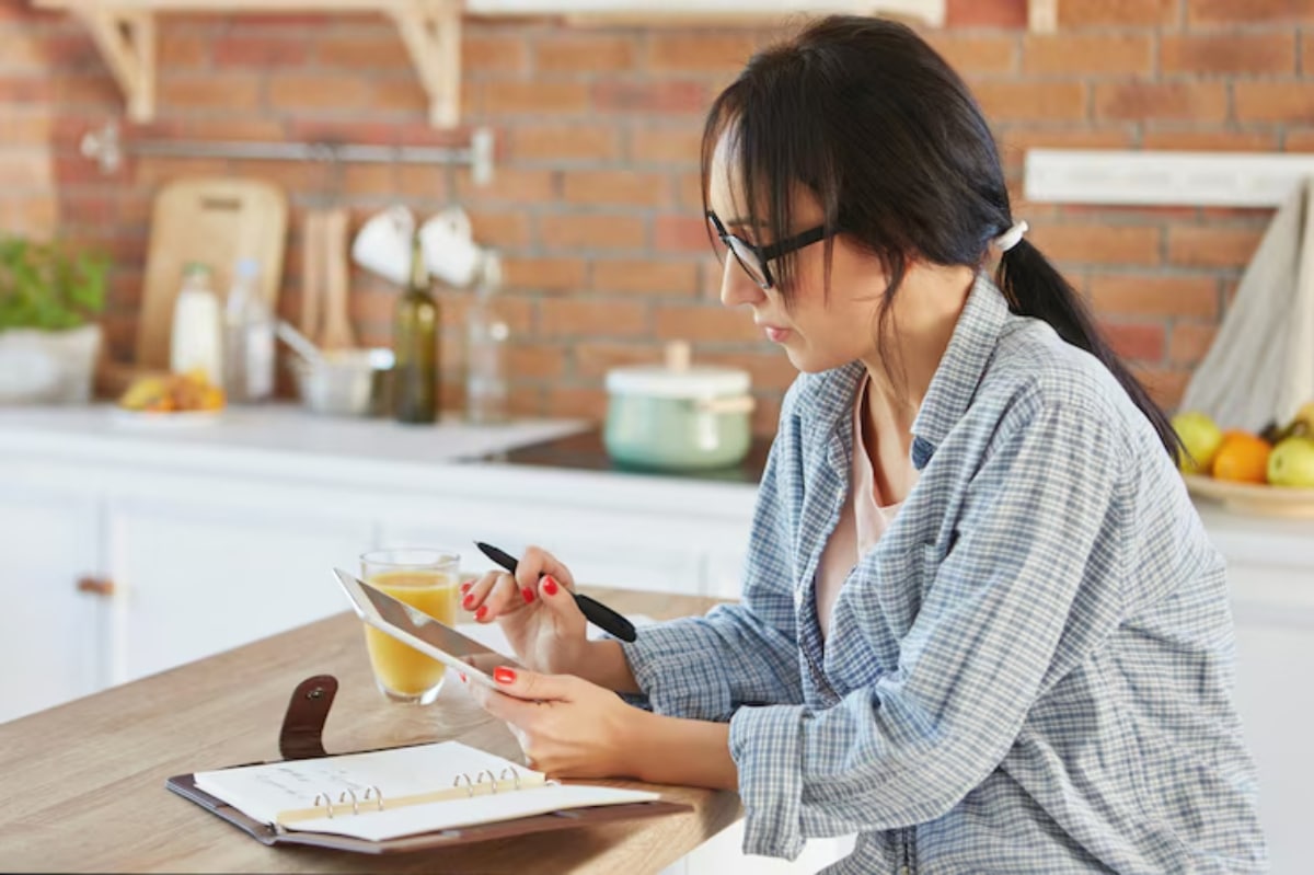 A person wearing a plaid shirt sits at a kitchen counter, holding a tablet and a pen, with a glass of juice nearby.