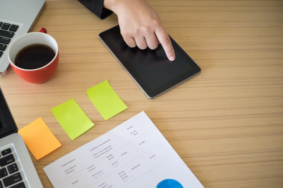 A hand points at a tablet on a wooden desk, alongside a laptop, sticky notes, a coffee cup, and a financial report.