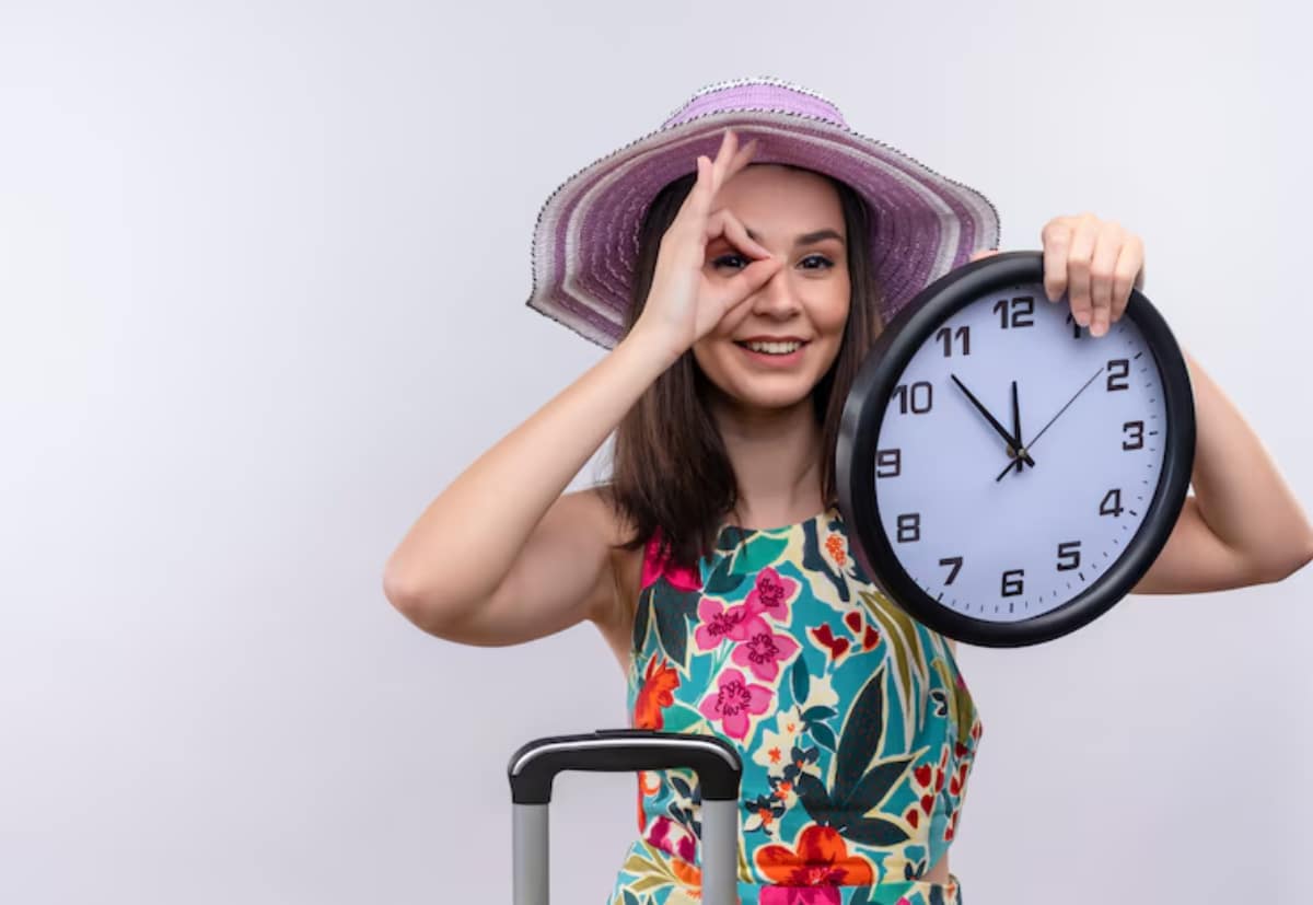 A woman in a floral dress and large hat holds a clock in one hand and touches her head with the other, standing beside a suitcase.