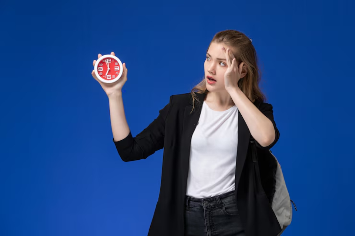 A woman in a black blazer and white shirt holds a round red clock against a bright blue background.
