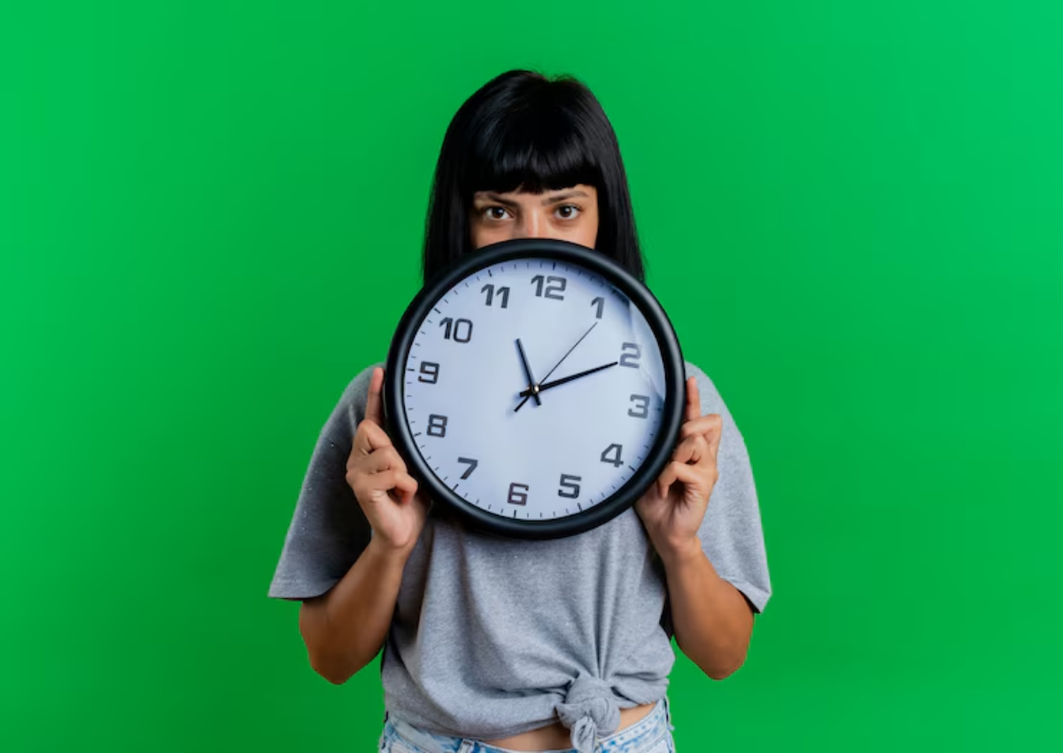 A person in a gray shirt holds a large clock against a bright green background. The clock shows the time clearly.