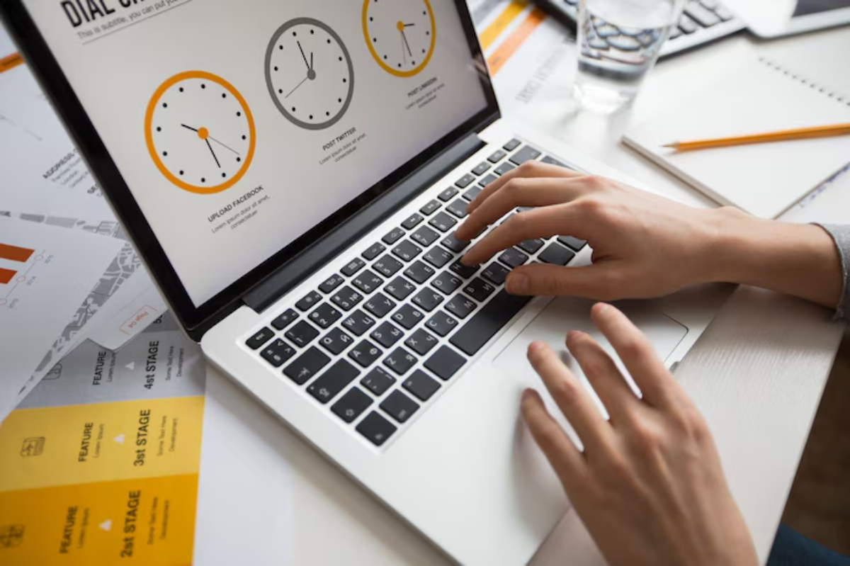 A person's hands typing on a laptop displaying a productivity dashboard with clock icons, surrounded by papers and a glass of water.