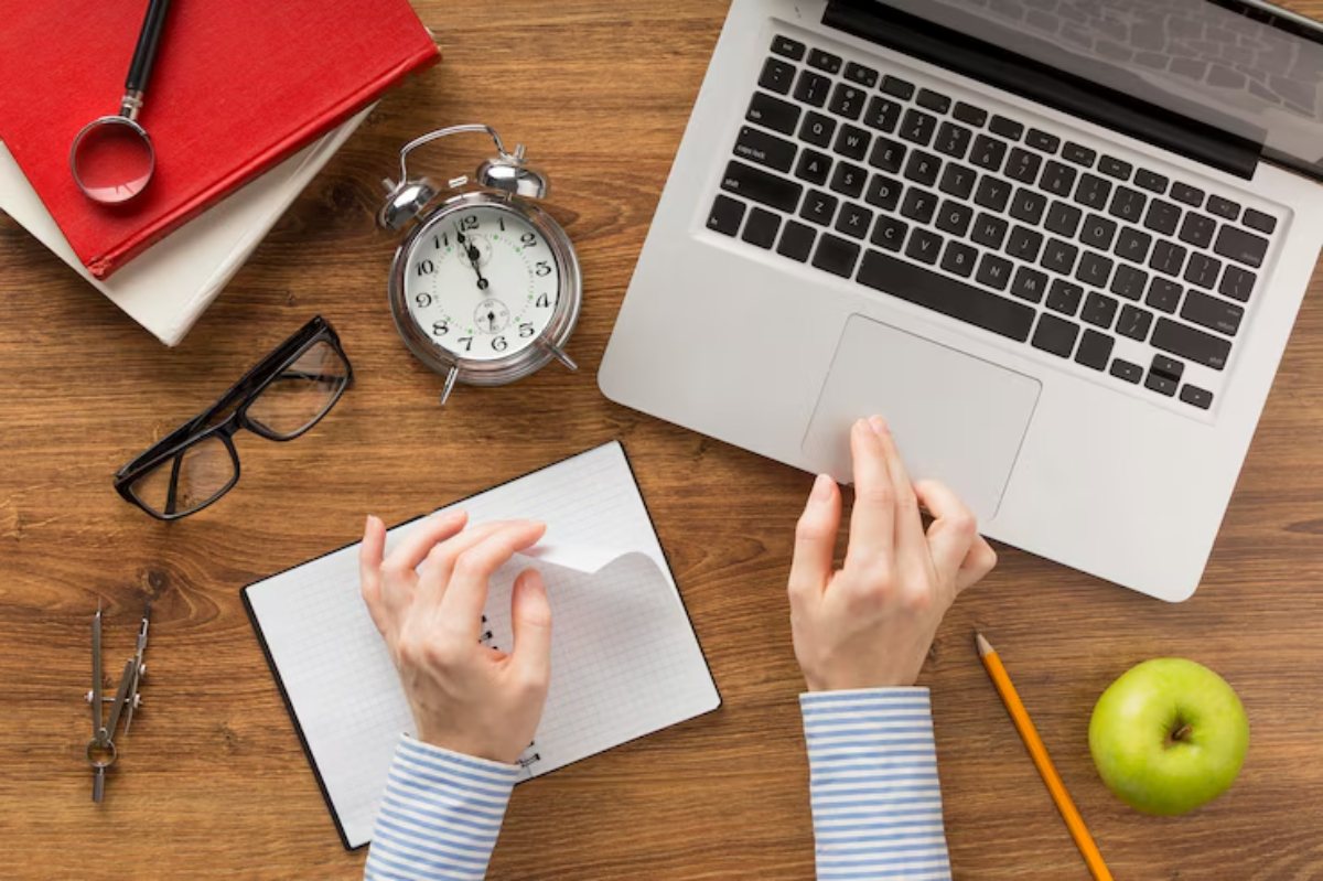 A top-down view of a workspace featuring a laptop, notebook, clock, apple, and glasses, with hands engaged in writing.