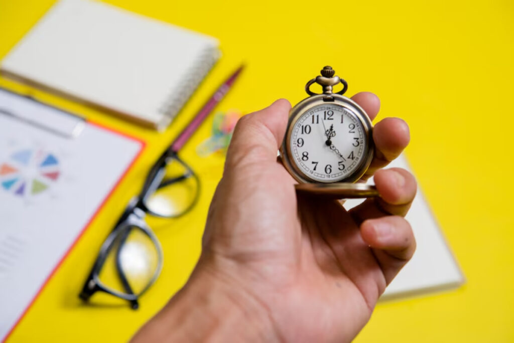 A hand holds a vintage pocket watch over a bright yellow table, surrounded by glasses, notebooks, and stationery.