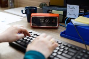 A person typing on a keyboard, with a vibrant orange clock displaying 12:26 and a blue coffee mug in the background.