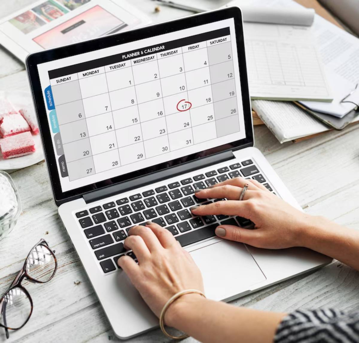 A person types on a laptop displaying a calendar, highlighting the 17th, surrounded by stationery and snacks on a wooden table. 